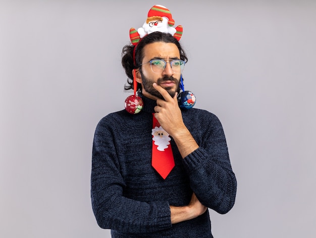 Denkender junger gutaussehender Kerl, der Weihnachtskrawatte mit Haarbügel hängenden Weihnachtsball auf Ohren packte Kinn lokalisiert auf weißem Hintergrund