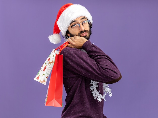 Denkender junger hübscher Kerl, der Weihnachtshut mit Girlande auf Hals hält, der Geschenktüten auf Schulter lokalisiert auf blauem Hintergrund hält