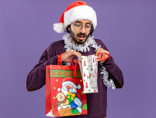 Denkender junger hübscher Kerl, der Weihnachtsmütze mit Girlande am Hals hält und in Geschenkbeutel lokalisiert auf blauem Hintergrund betrachtet