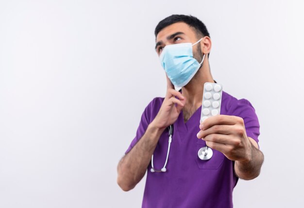 Denkender junger männlicher Arzt, der lila Chirurgenkleidung und medizinische Stethoskopmaske trägt, die Pillen auf isoliertem Weiß heraushalten