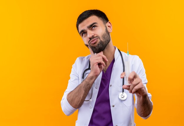 Denkender junger männlicher Arzt, der medizinisches Kleid des Stethoskops hält Spritze auf isolierter gelber Wand hält