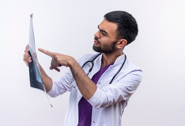 Denkender junger männlicher Arzt, der medizinisches Kleid des Stethoskops trägt, zeigt auf Röntgen in seiner Hand auf lokalisiertem Weiß