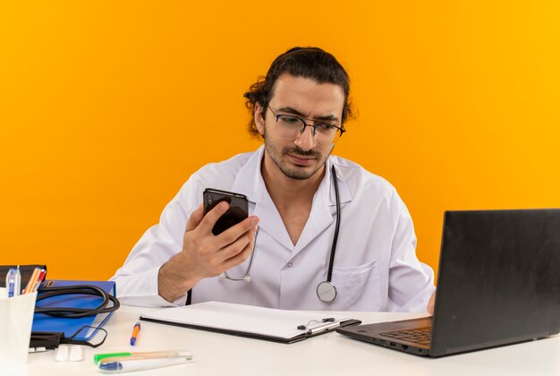 Denkender junger männlicher Arzt mit medizinischer Brille mit medizinischem Gewand mit Stethoskop am Schreibtisch