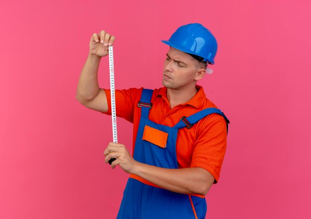 Denkender junger männlicher Baumeister, der Uniform und Schutzhelm trägt, der Meterband auf Rosa hält und betrachtet