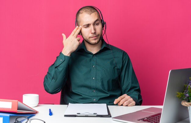 Denkender junger männlicher Callcenter-Betreiber, der ein Headset am Schreibtisch mit Bürowerkzeugen trägt und auf den Laptop schaut, der Selbstmordgeste mit Pistolengeste zeigt