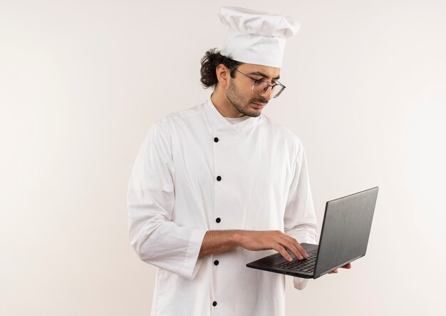 Denkender junger männlicher Koch, der Kochuniform und Brille hält und gebrauchten Laptop lokalisiert auf weißer Wand trägt