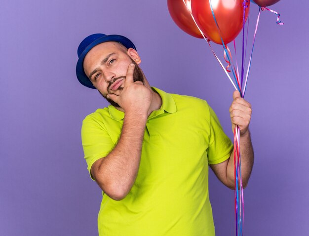 Denkender junger Mann mit Partyhut, der Luftballons hält und das Kinn packte