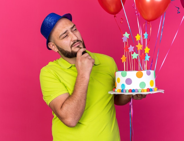 Denkender junger Mann mit Partyhut isoliert auf Luftballons mit Kuchen packte das Kinn isoliert auf rosa Wand
