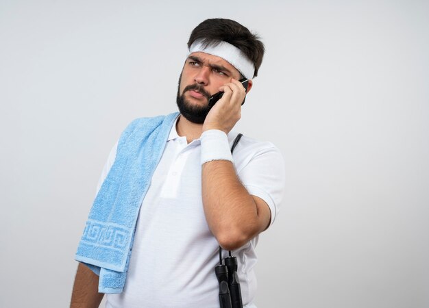 Denkender junger sportlicher Mann, der Seite betrachtet, die Stirnband und Armband mit Handtuch und Springseil auf Schulter trägt, spricht am Telefon lokalisiert auf weißer Wand mit Kopienraum