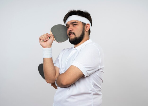 Denkender junger sportlicher Mann, der Seite betrachtet, die Stirnband und Armband trägt, die Tischtennisschläger um Gesicht lokalisiert auf weißer Wand halten