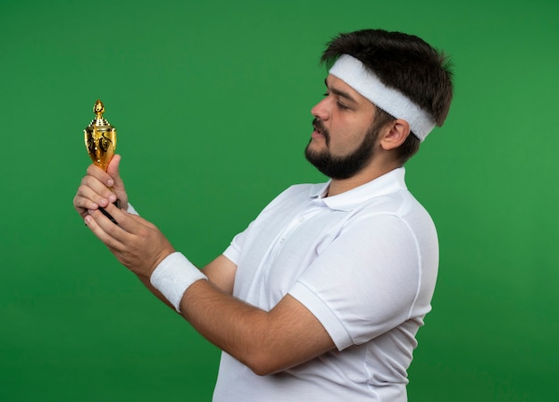 Denkender junger sportlicher Mann, der Stirnband und Armband hält und Siegerpokal lokalisiert auf grüner Wand hält