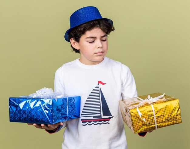 Denkender kleiner Junge mit blauem Partyhut, der Geschenkboxen hält und betrachtet