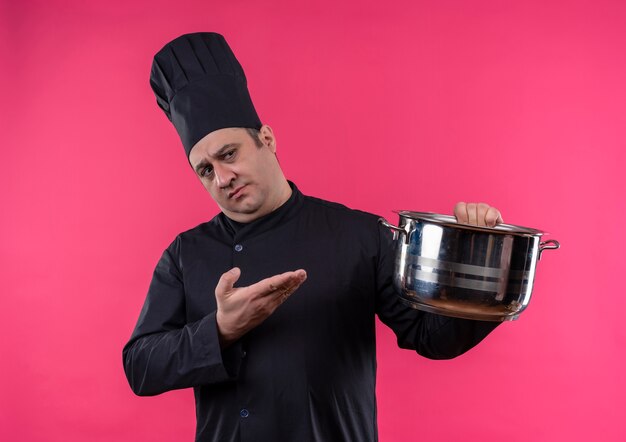 Denkender männlicher Koch mittleren Alters in der Kochuniform, die Topf in seiner Hand zeigt