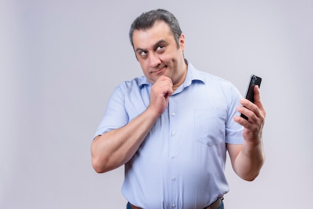 Denkender Mann mittleren Alters, der blaues gestreiftes Hemd hält, das sein Handy mit Hand hält, während auf einem weißen Hintergrund stehend