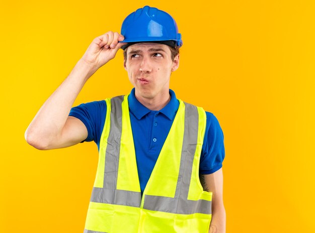 Denkender seitlicher junger Baumeister in Uniform isoliert auf gelber Wand