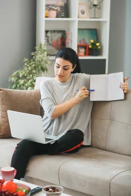 Denkendes junges Mädchen benutzte Laptop-Haltung und Punkte auf Buch mit Stift, der auf Sofa im Wohnzimmer sitzt