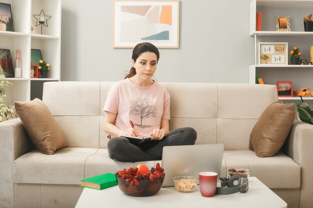 Denkendes junges Mädchen, das ein Notebook auf dem Sofa hinter dem Couchtisch hält und einen Laptop im Wohnzimmer betrachtet