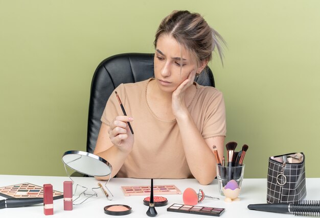 Denkendes junges schönes Mädchen, das am Schreibtisch mit Make-up-Tools sitzt und Make-up-Pinsel hält und betrachtet, die Hand auf die Wange legt, einzeln auf olivgrünem Hintergrund