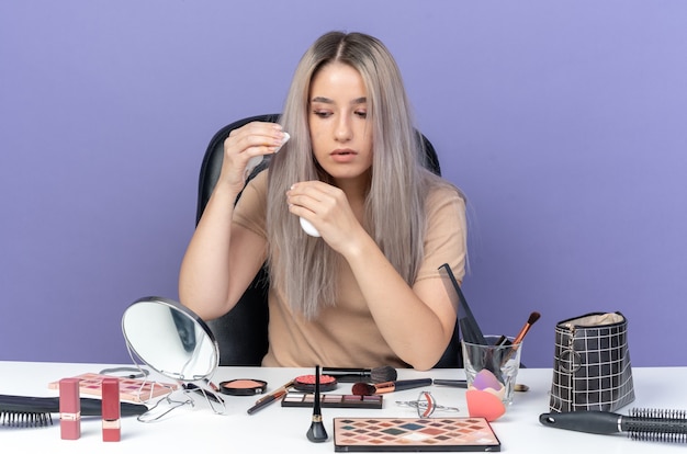 Denkendes junges schönes Mädchen sitzt am Tisch mit Make-up-Tools und hält Haarcreme isoliert auf blauem Hintergrund