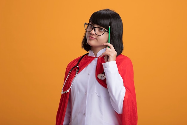 Denkendes junges Superheldenmädchen, das Stethoskop mit medizinischem Gewand und Umhang mit Brille trägt, die Kopf mit Bleistift kratzt