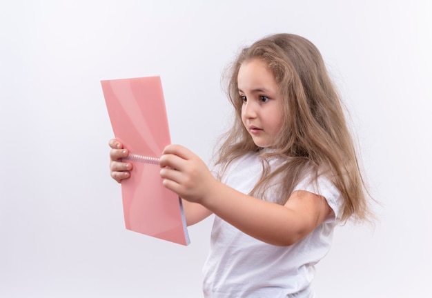 Denkendes kleines Schulmädchen, das weißes T-Shirt trägt, das Notizbuch auf isolierter weißer Wand betrachtet