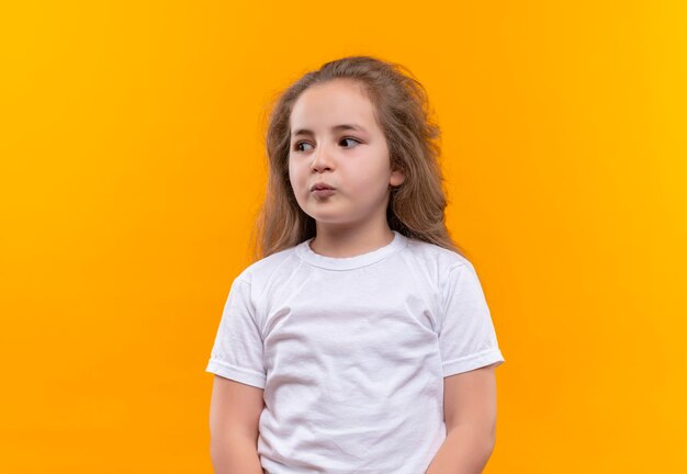Denkendes kleines Schulmädchen, das weißes T-Shirt trägt, das Seite auf isolierte orange Wand betrachtet