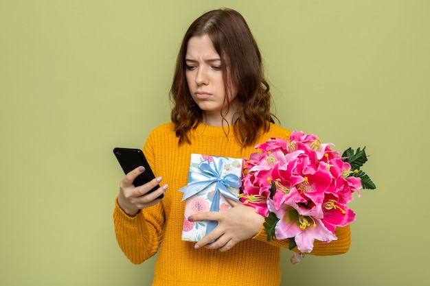 Denkendes schönes junges Mädchen am Tag der glücklichen Frau, das Geschenke hält und das Telefon in der Hand betrachtet, das auf olivgrüner Wand isoliert ist?