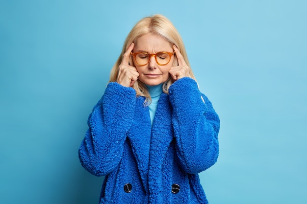 Depressive blonde vierzig Jahre alte Frau hält Finger an den Schläfen denkt tief oder hat Kopfschmerzen schließt die Augen, als versucht, sich zu konzentrieren, trägt Wintermantel.