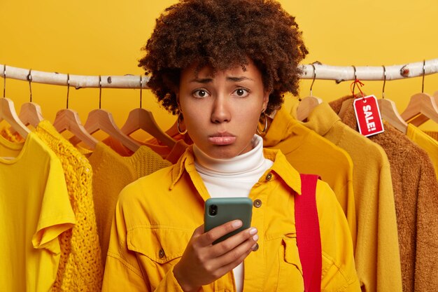 Deprimierte Afro-Frau nutzt Smartphone zum Online-Shopping, unglücklich, steht gegen gelbe Kleidung in Kleiderbügeln