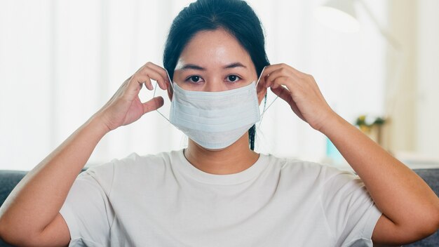 Deprimierte asiatische Geschäftsfrau, die Schutzmaske trägt, die auf Sofa im Wohnzimmer am Haus sitzt, wenn soziale Distanzierung zu Hause bleibt und Selbstquarantänezeit, Pandemie in China, Coronavirus-Konzept.