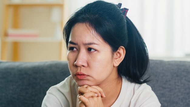 Deprimierte weinende asiatische Frau gestresst mit Kopfschmerzen, die auf Sofa im Wohnzimmer am Haus sitzen