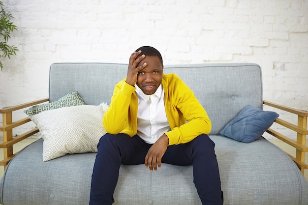 Deprimierter, lässig gekleideter junger afroamerikaner, der zu hause auf der couch sitzt, die hand auf dem kopf hält, die fußballmeisterschaft beobachtet und sich verärgert fühlt, während seine lieblingsmannschaft das spiel verliert