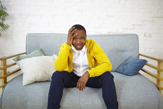 Deprimierter, lässig gekleideter junger Afroamerikaner, der zu Hause auf der Couch sitzt, die Hand auf dem Kopf hält, die Fußballmeisterschaft beobachtet und sich verärgert fühlt, während seine Lieblingsmannschaft das Spiel verliert