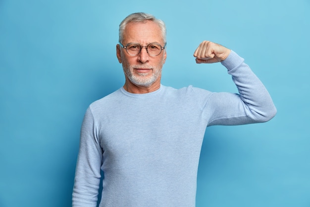 Der ältere bärtige Mann zeigt Muskeln, nachdem er Bodybuilding geübt hat. Er trägt eine transparente Brille und einfache Pullover-Posen gegen die blaue Studiowand