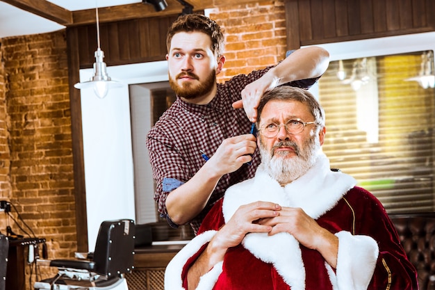 Der ältere Mann im Weihnachtsmannkostüm rasiert seinen persönlichen Meister vor Weihnachten im Friseurladen