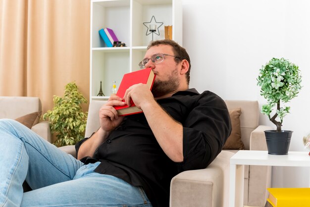 Der ängstliche erwachsene slawische Mann in der optischen Brille sitzt auf dem Sessel, der Buch nahe am Mund hält und im Wohnzimmer nach oben schaut