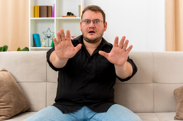 Der ängstliche erwachsene slawische Mann in der optischen Brille sitzt auf dem Sessel und streckt die Hände im Wohnzimmer aus