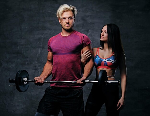 Der athletische, blonde Mann hält Langhantelgewicht und schlankes Fitness-Frauenmodell vor dunkelgrauem Hintergrund.