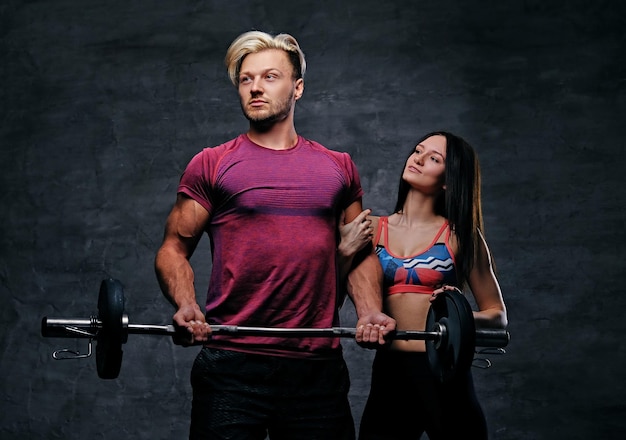 Der athletische, blonde Mann hält Langhantelgewicht und schlankes Fitness-Frauenmodell vor dunkelgrauem Hintergrund.