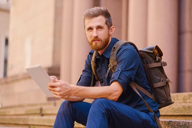 Der bärtige Gelegenheitsreisende sitzt auf einer Stufe und benutzt einen Tablet-PC.