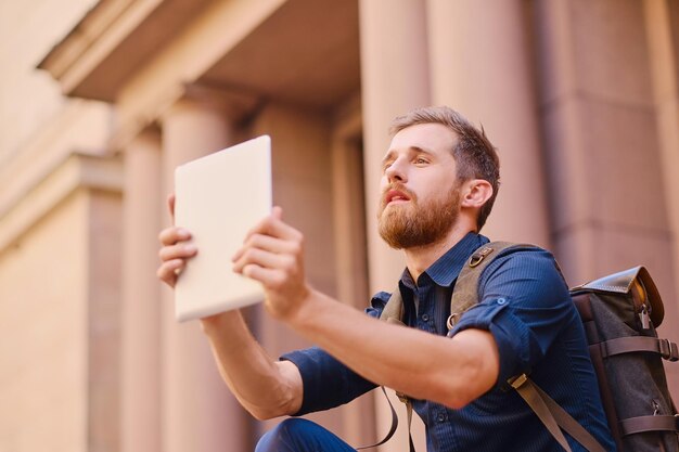 Der bärtige Gelegenheitsreisende sitzt auf einer Stufe und benutzt einen Tablet-PC.