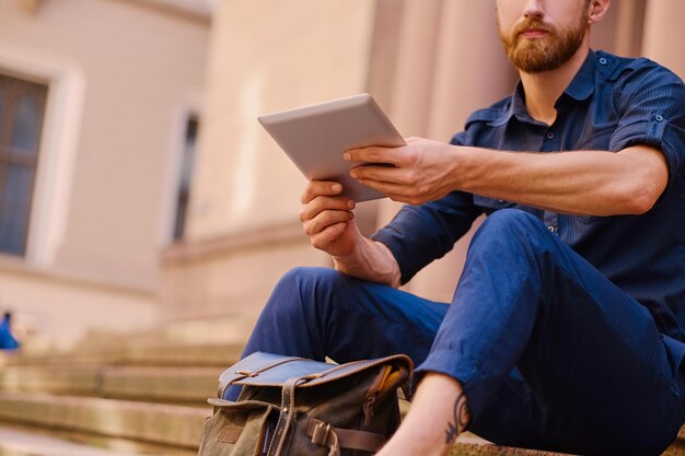 Der bärtige Gelegenheitsreisende sitzt auf einer Stufe und benutzt einen Tablet-PC.