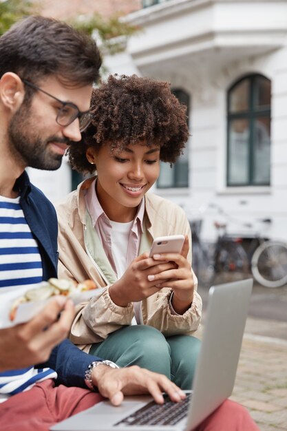 Der bärtige Hipster-Typ durchsucht die Website nach Netzwerken und isst Junk Food. Afroamerikanisches Mädchen benutzt ein Handy