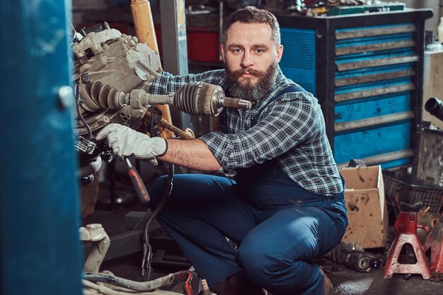 Der bärtige Mechaniker repariert den Automotor, der auf der hydraulischen Hebebühne in der Garage angehoben wird. Tankstelle.