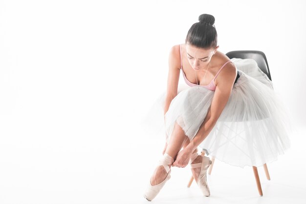 Der Balletttänzer, der auf Stuhl sitzt, binden oben ihre pointe Schuhe gegen weißen Hintergrund