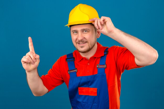 Der Baumeister, der eine Bauuniform und einen Sicherheitshelm trug, hatte die Idee, mit dem Finger nach oben zu zeigen und seinen Helm zu berühren, der über einer isolierten blauen Wand stand