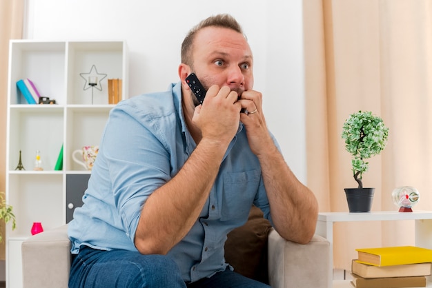 Der besorgte erwachsene slawische Mann sitzt auf dem Sessel und hält die fernbeißenden Finger des Fernsehers und schaut auf die Seite im Wohnzimmer