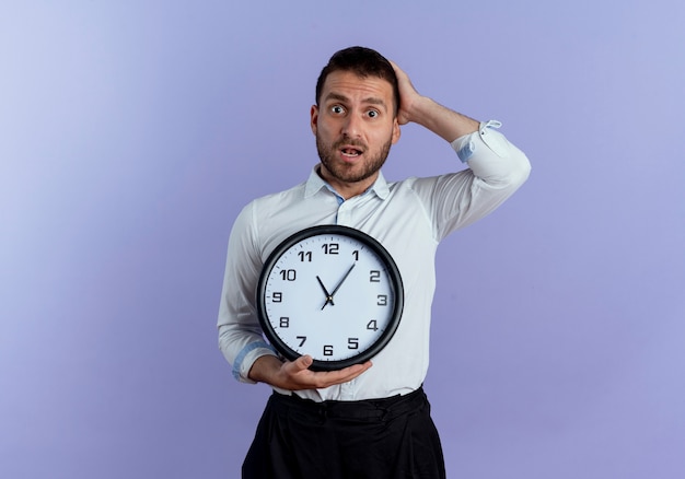 Der besorgte gutaussehende Mann legt die Hand auf den Kopf hinter die Uhr, die auf lila Wand isoliert hält