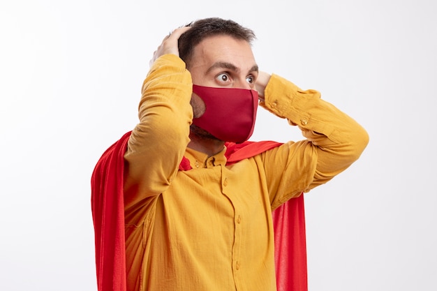 Der besorgte Superheldenmann mit dem roten Umhang, der rote Maske trägt, setzt Hände auf Kopf, der Seite betrachtet, die auf weißer Wand lokalisiert wird