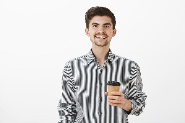 Der charmante, freundliche Kerl brachte dem Kollegen, den er mag, Kaffee. Porträt des erfreuten niedlichen Freundes im Hemd, breit lächelnd und Tasse Tee haltend, beiläufig mit Kollegen im Büro über graue Wand sprechend